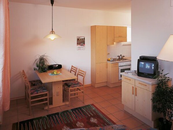 A compact kitchen with wooden cabinets, a dining table with chairs, a TV, and a hanging light fixture. - Appartment Mühlrast (Laces)
