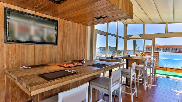 Una zona de bar con barra de madera, un televisor grande, sillas blancas y vista al mar a través de amplias ventanas.