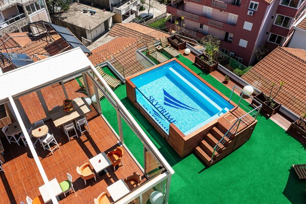 Un espace piscine sur le toit, avec une piscine bleue, une pelouse artificielle verte et des sièges en bois sur une terrasse.