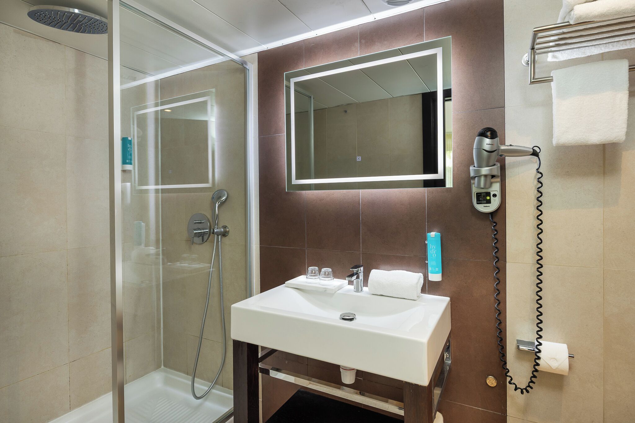 Een moderne badkamer met een glazen douchecabine, een wit wastafeltje met een spiegel erboven en een haardroger aan de muur.