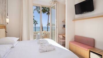 A hotel room with a bed, a desk, a pink chair, a TV, and a view of the sea.