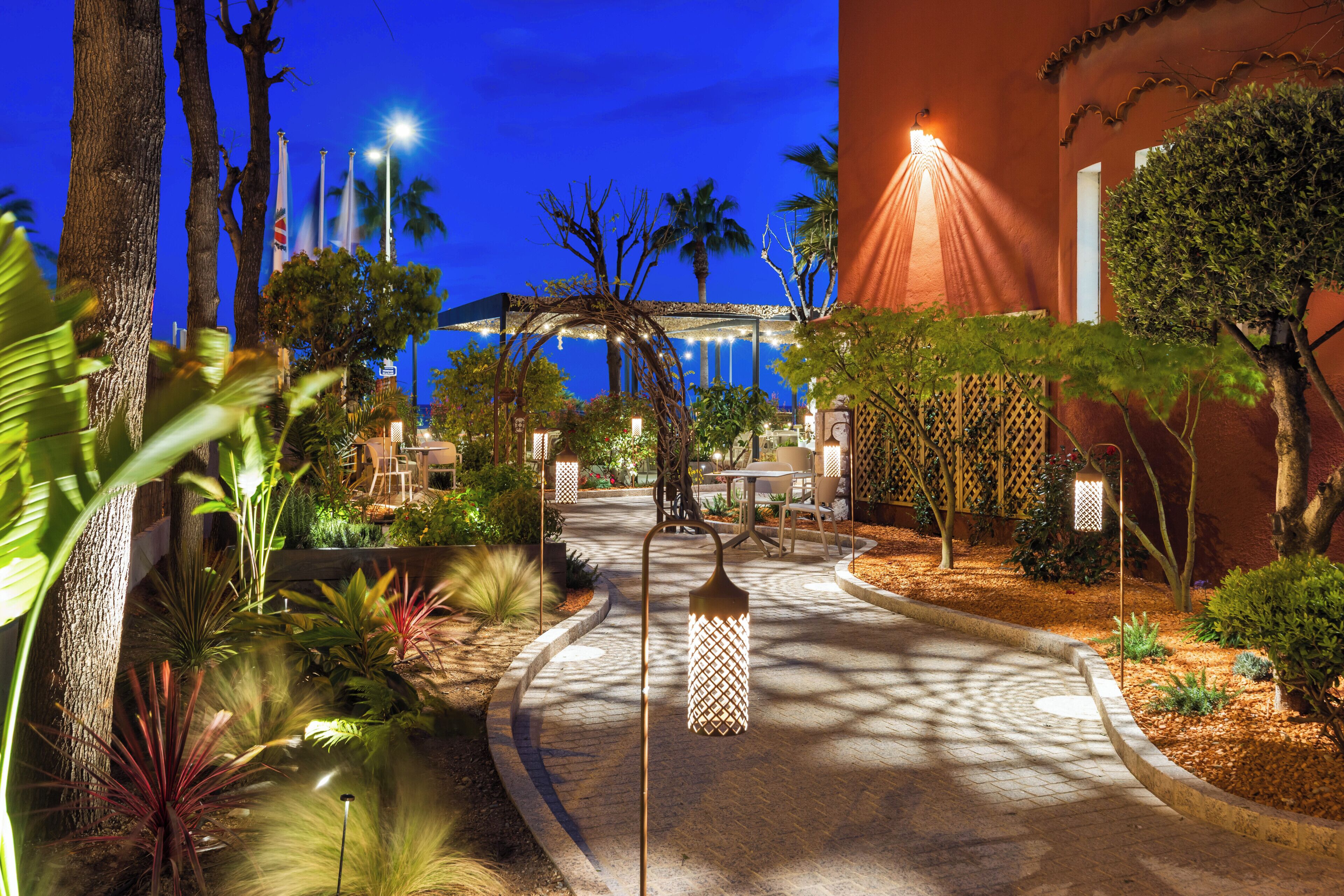 A well-lit outdoor area with a curved pathway, palm trees, and a building with a red facade.