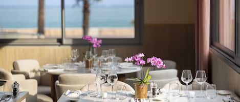 A dining area with tables set for two, featuring a view of the ocean through large windows.