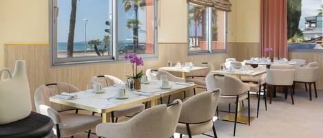 A restaurant with multiple tables and chairs, large windows with curtains, and a view of palm trees and the sea.
