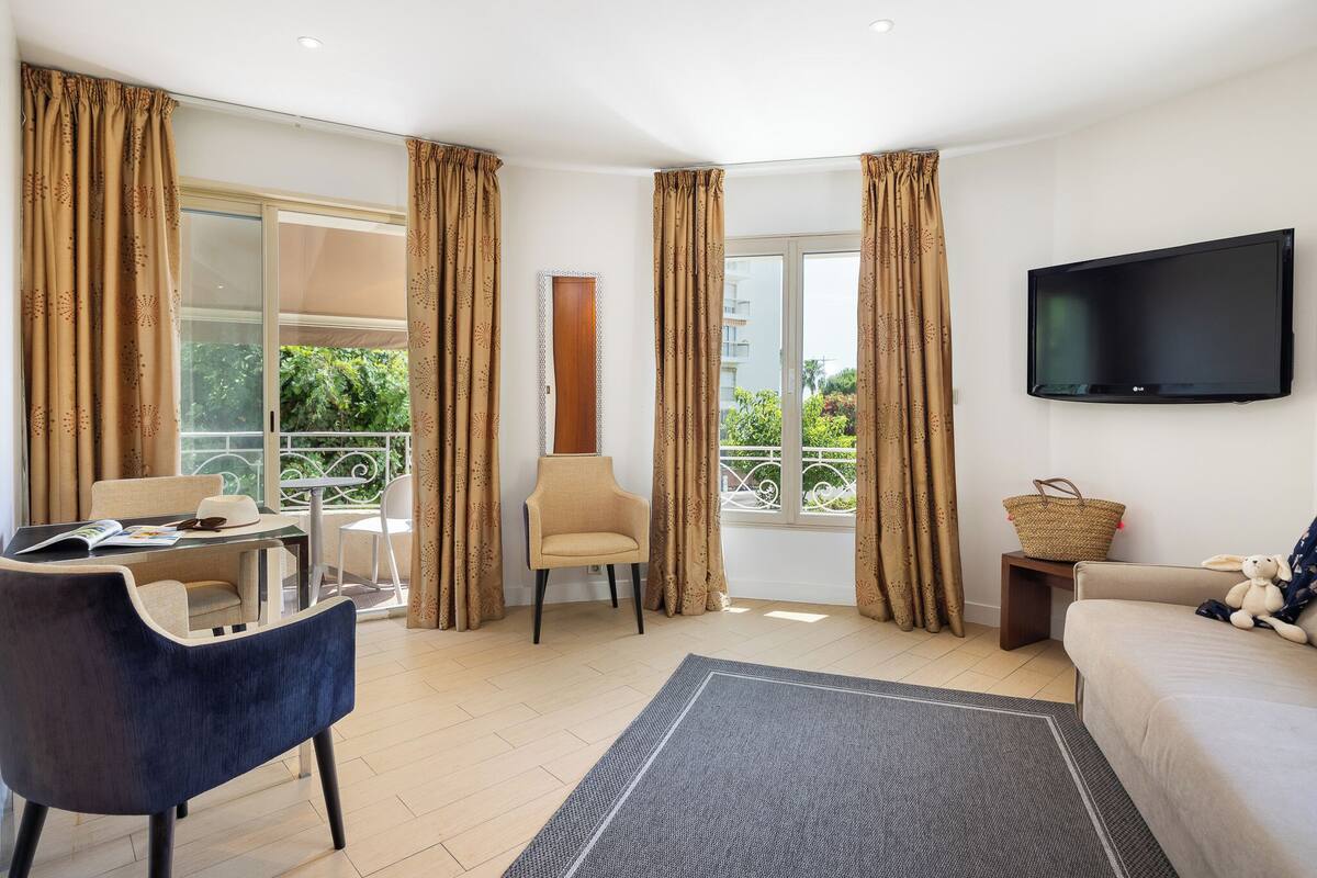 Un salon moderne avec un canapé, un fauteuil et une table à manger avec des chaises. Un téléviseur est fixé au mur et un balcon est visible à travers la fenêtre.