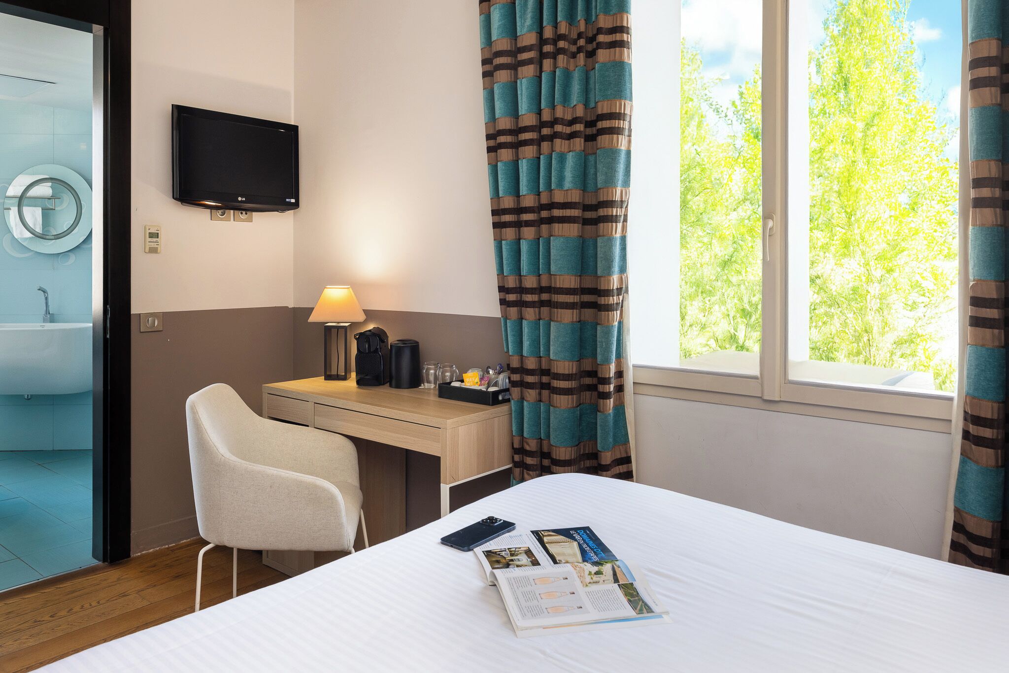 Ein Hotelzimmer mit Bett, Schreibtisch, Stuhl, Fernseher und einem Fenster mit Vorhängen.