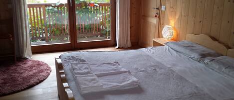 Un dormitorio con cama, cabecera de madera, mesita de noche con lámpara, ventana con vista a una valla de madera y follaje, y una alfombra mullida.