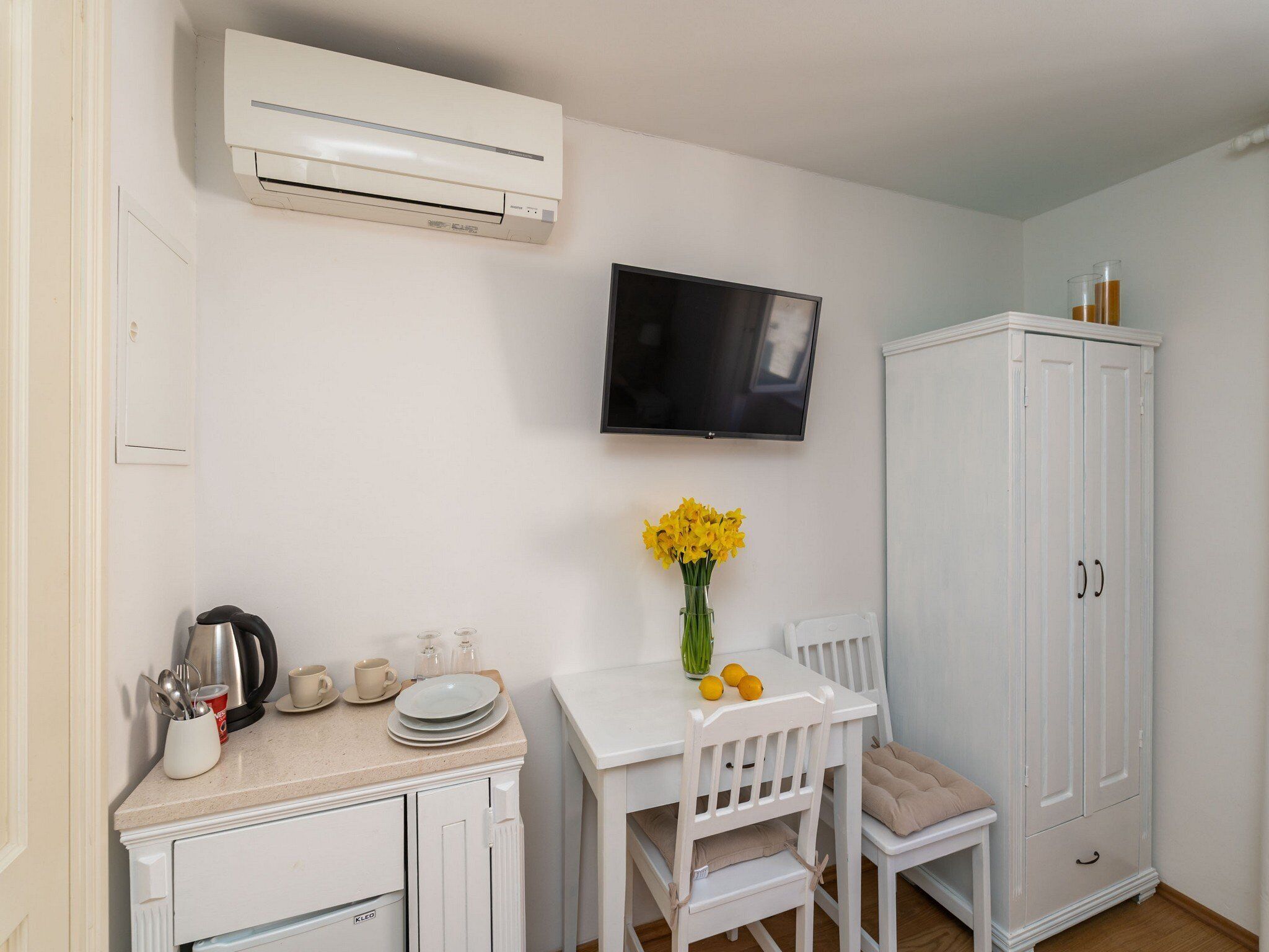 A compact kitchen with white cabinetry, a white dining table with chairs, a mounted flat-screen TV, and a white cabinet with a mirror.
