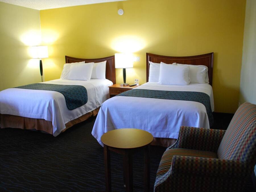 Ein Hotelzimmer mit zwei Betten, jeweils mit einem hölzernen Kopfteil, weißer Bettwäsche und einer gemusterten Tagesdecke. Es gibt einen kleinen runden Tisch und einen gemusterten Sessel.