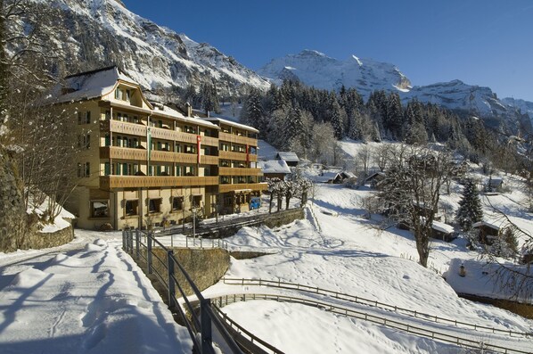 Ein mehrstöckiges Hotel mit Balkonen, umgeben von schneebedeckten Bergen und Bäumen.