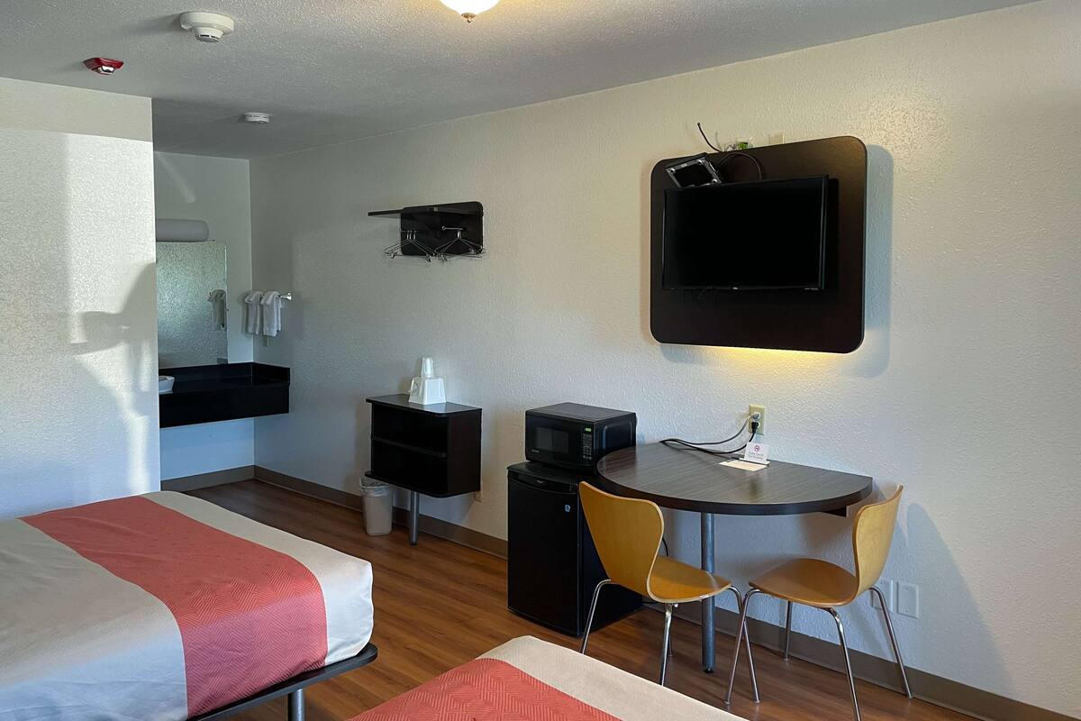 Habitación de hotel con dos camas, un escritorio con silla, microondas, televisor y un estante pequeño con artículos de aseo.
