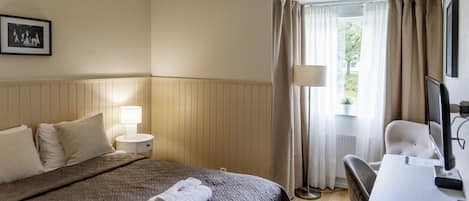 Una habitación de hotel con cama, escritorio, silla, lámpara y ventana con cortinas.