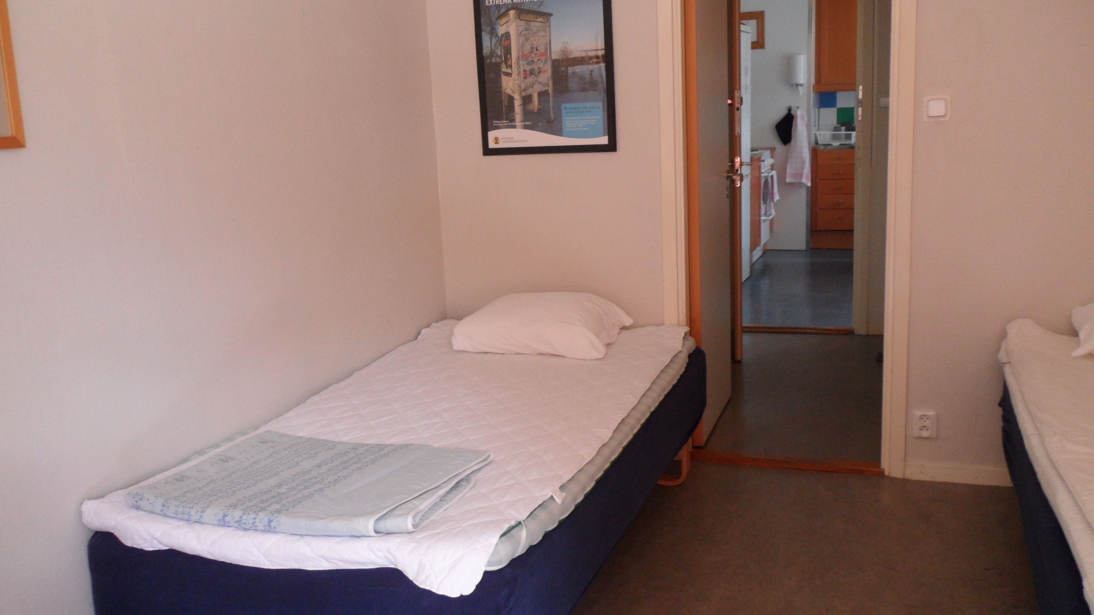 Een kleine kamer met een enkel bed, een effen witte muur, een deur naar een badkamer en een klein raam.