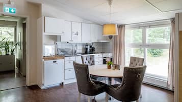 Ett modernt kök med vita skåp, ett matbord med stolar, en hängande lampa och utsikt över grönområden utanför.