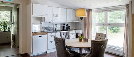 Une cuisine moderne avec des meubles blancs, une table à manger avec des chaises, une lampe suspendue et une vue sur la verdure à l’extérieur.