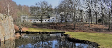 Een groot, historisch gebouw met meerdere verdiepingen en een opvallende toren, omgeven door een verzorgd gazon en een kleine vijver die de structuur weerspiegelt.