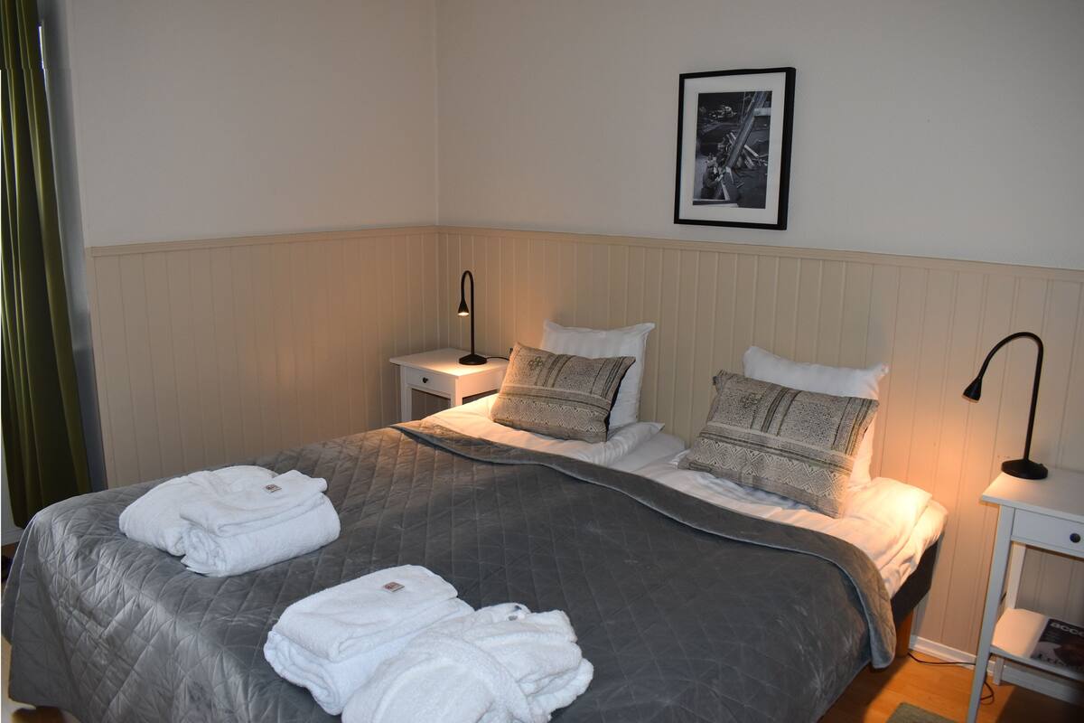 Een keurig opgemaakt bed met een grijze deken, twee witte handdoeken en decoratieve kussens. Daarboven hangt een lijst met een afbeelding aan de muur.