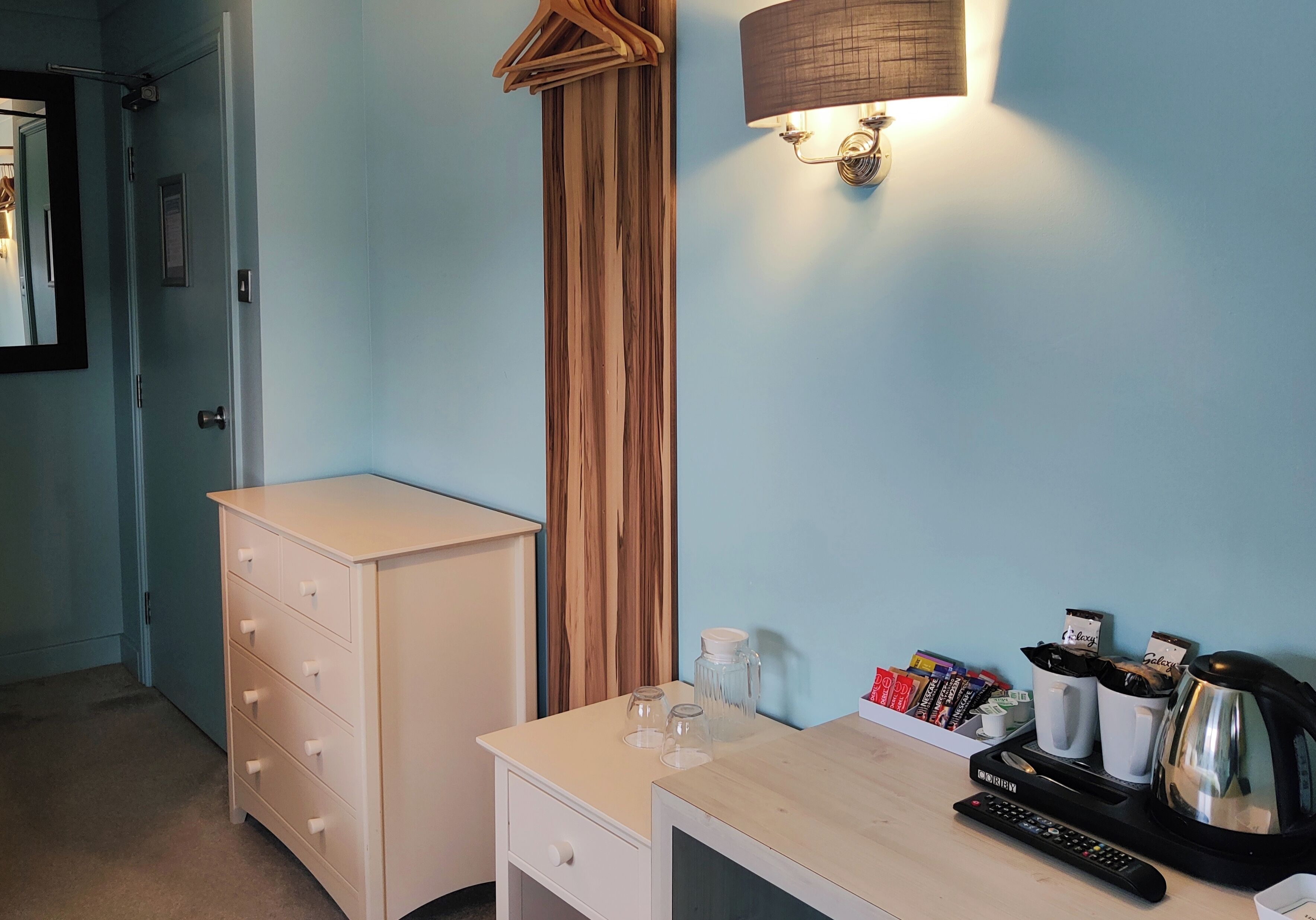 A neatly organized room with a dresser, a bedside table with a kettle, cups, and a remote control.