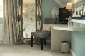 A room with a grey chair, a small desk with a lamp, a TV, a fan, and a trash bin.