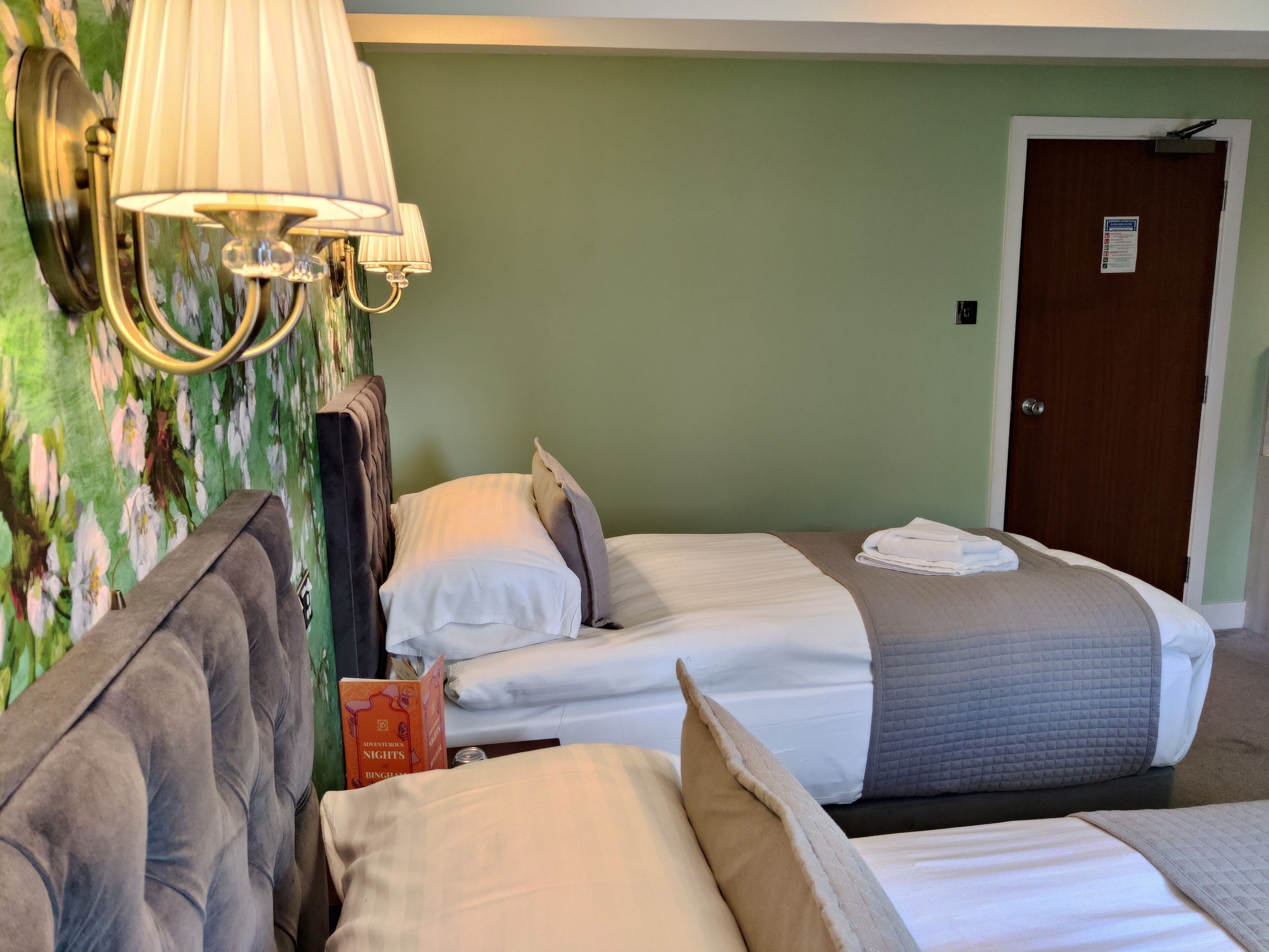 Una habitación de hotel con dos camas, una mesita de noche, una lámpara y un papel tapiz con motivos florales.