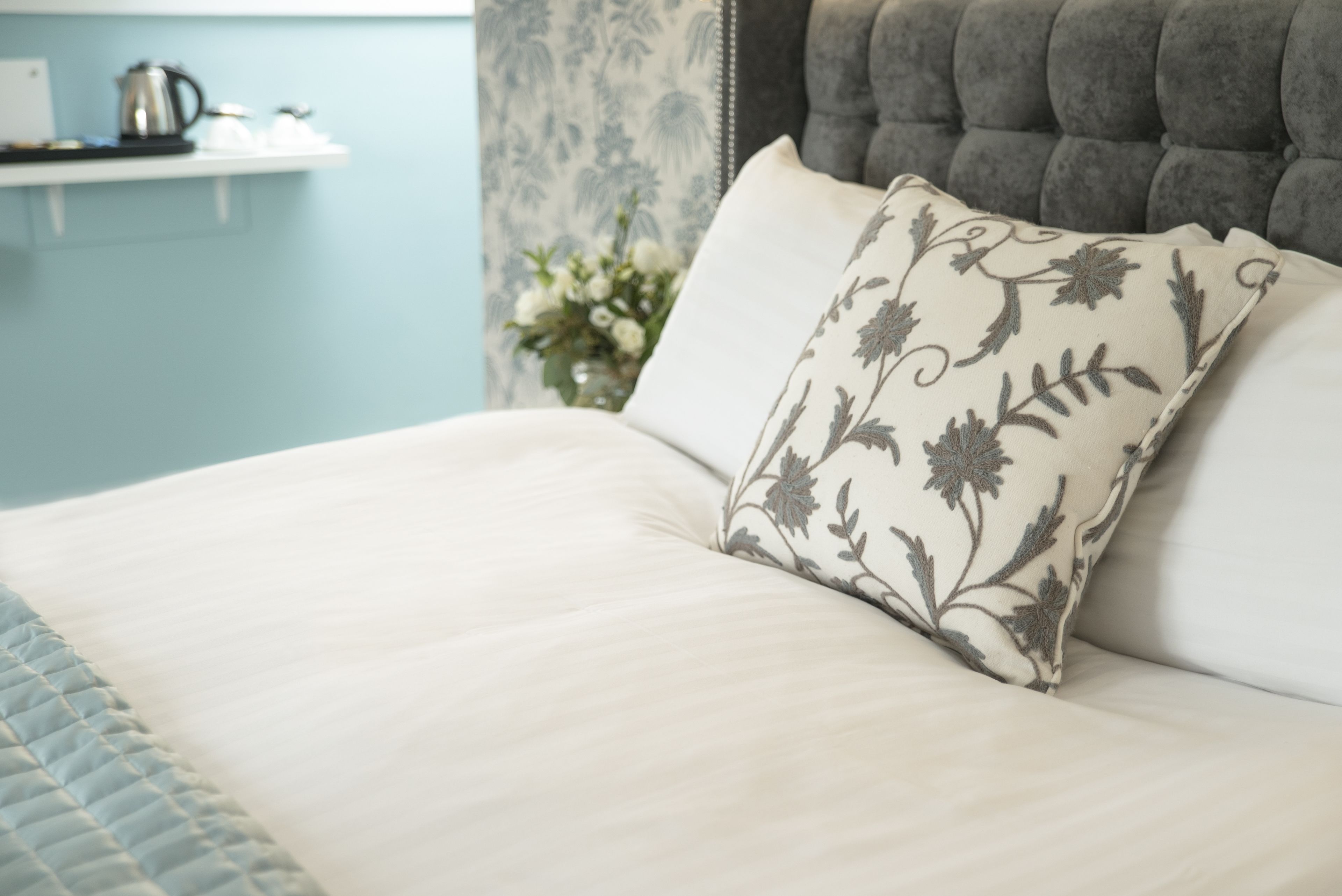 A bed with a floral pillow, a tufted headboard, and a light blue wall with a shelf holding a kettle and cups.