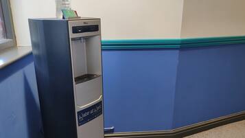 Un dispensador de agua con una botella de gel antibacterial encima, un cartel que dice 'Agua para el Trabajo' y una pared azul con detalles color turquesa.