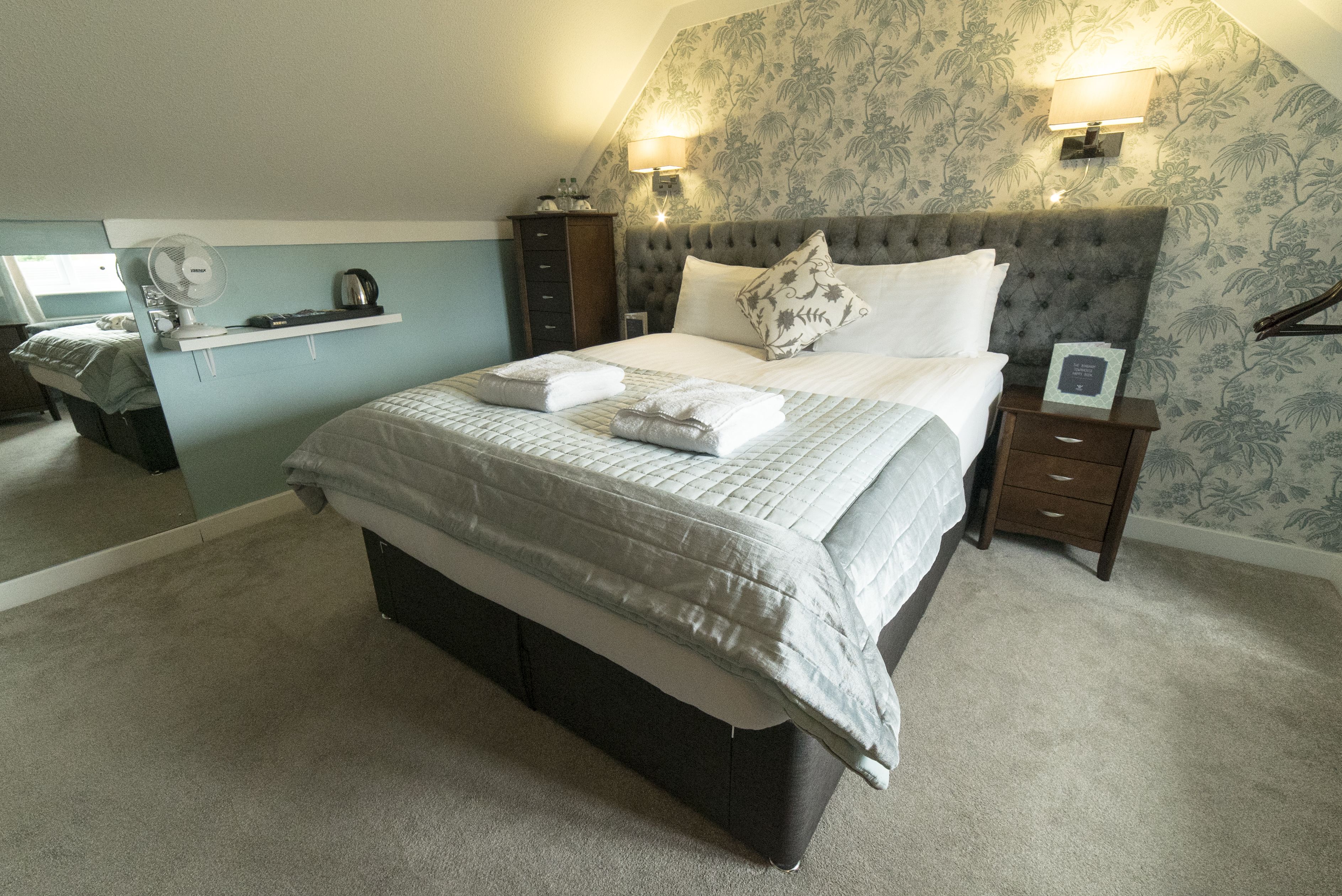 A neatly made bed with a grey and white duvet, a bedside table with a lamp, and a nightstand with a framed picture.