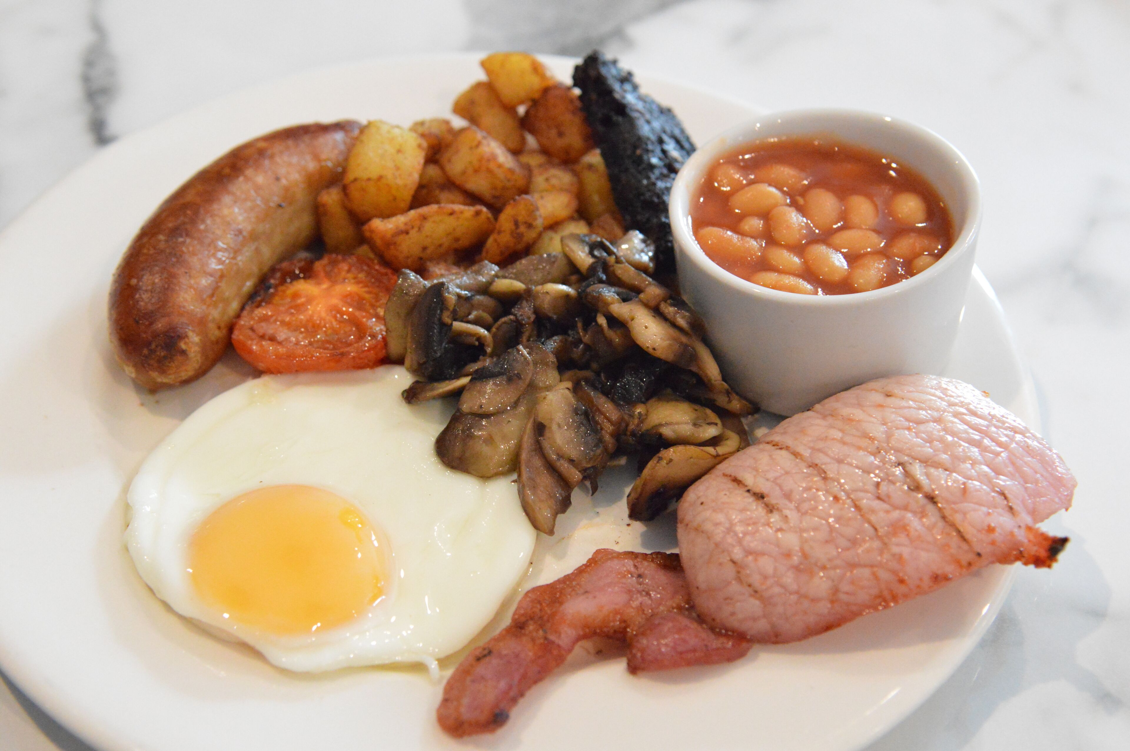 A plate of breakfast food including eggs, bacon, sausages, mushrooms, baked beans, and roasted potatoes.