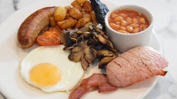 A plate of breakfast food including eggs, bacon, sausages, mushrooms, baked beans, and roasted potatoes.