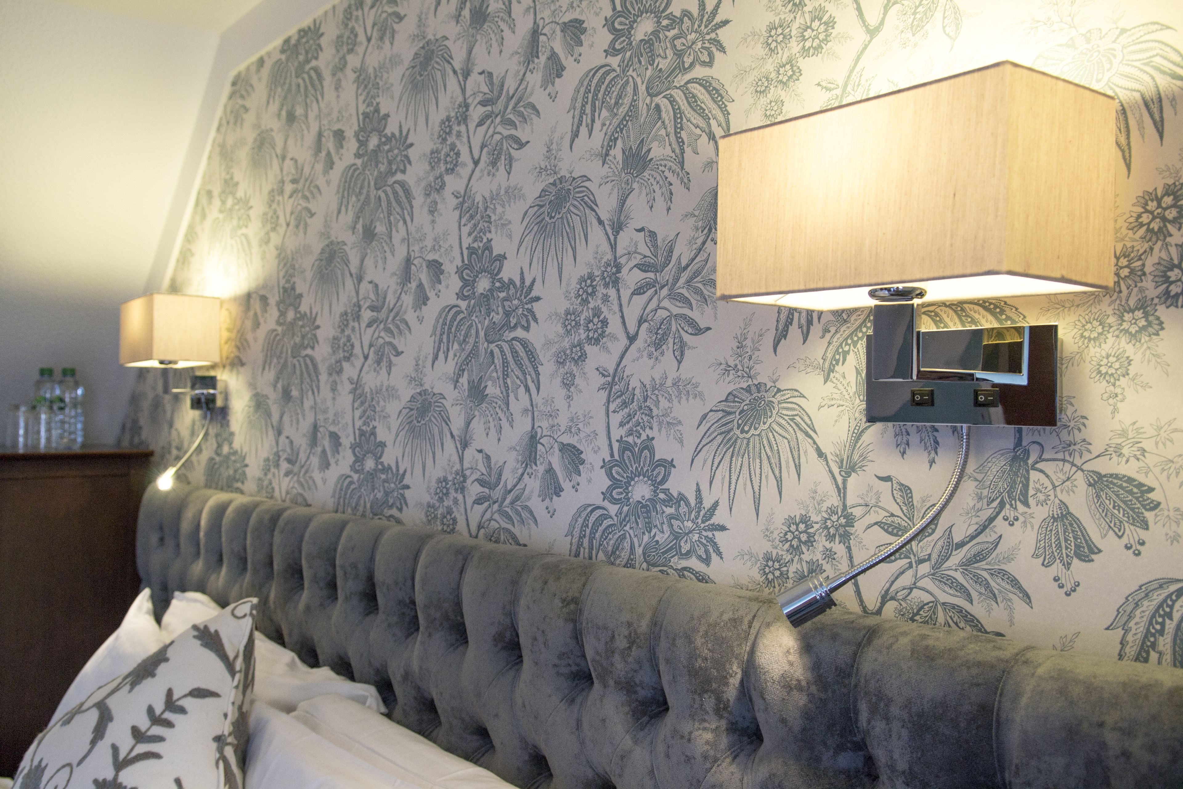 A bedroom with a tufted headboard, floral wallpaper, and two wall-mounted lamps.