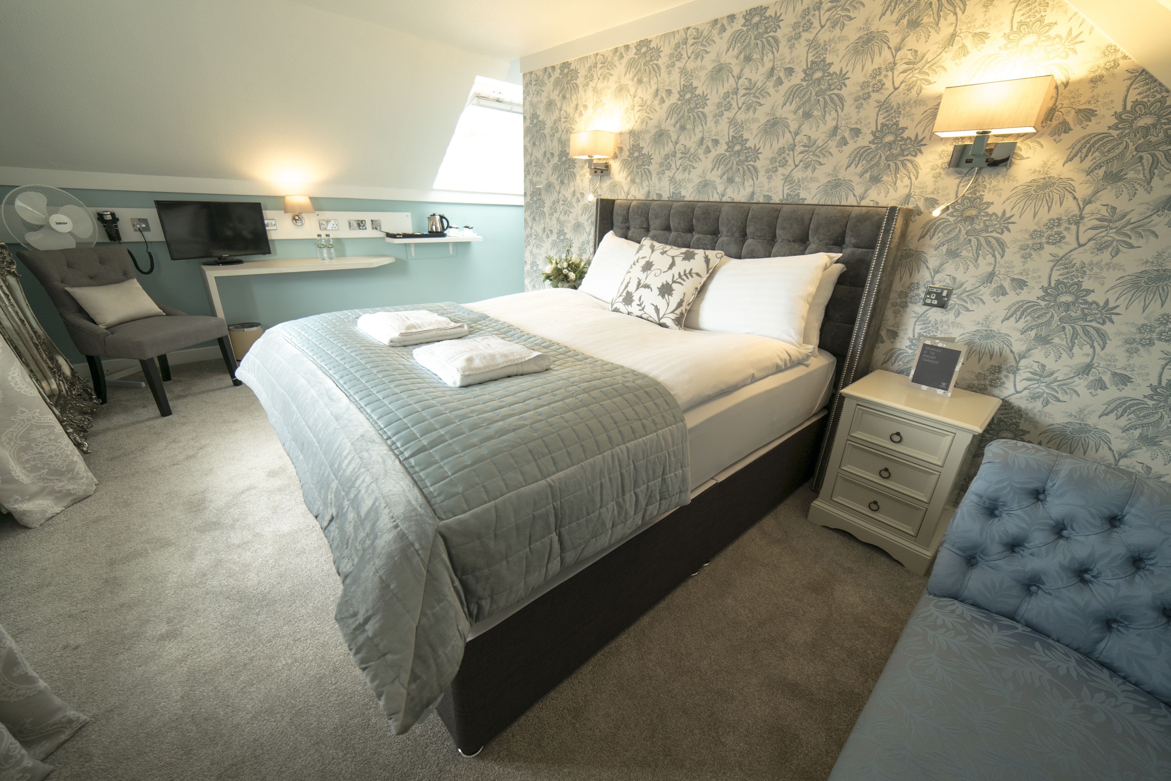 A bedroom with a bed, bedside tables, a chair, a desk, a TV, and a wall-mounted lamp.