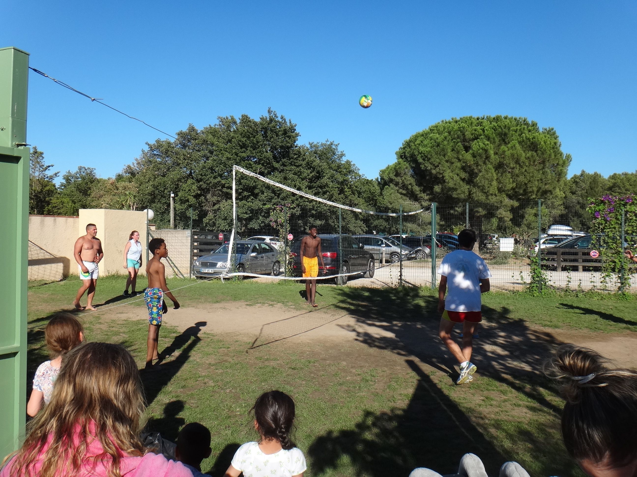 A group of people playing volleyball in an outdoor setting with a net, trees, and parked cars in the background.