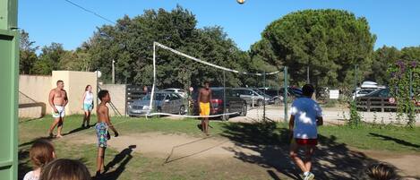 Un grupo de personas jugando voleibol al aire libre, con una red, árboles y coches estacionados al fondo.