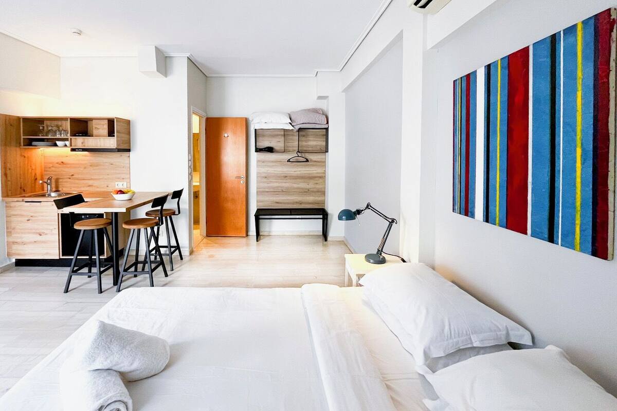 A modern hotel room with a large bed, a desk with chairs, a small kitchen area, and a colorful abstract painting on the wall.