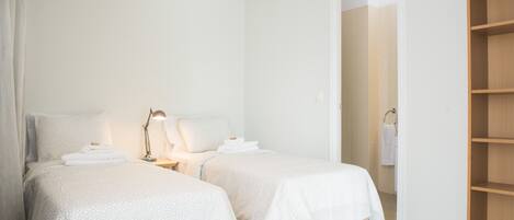 A clean, modern bedroom with two beds, a bedside table with a lamp, and a wooden shelf unit.
