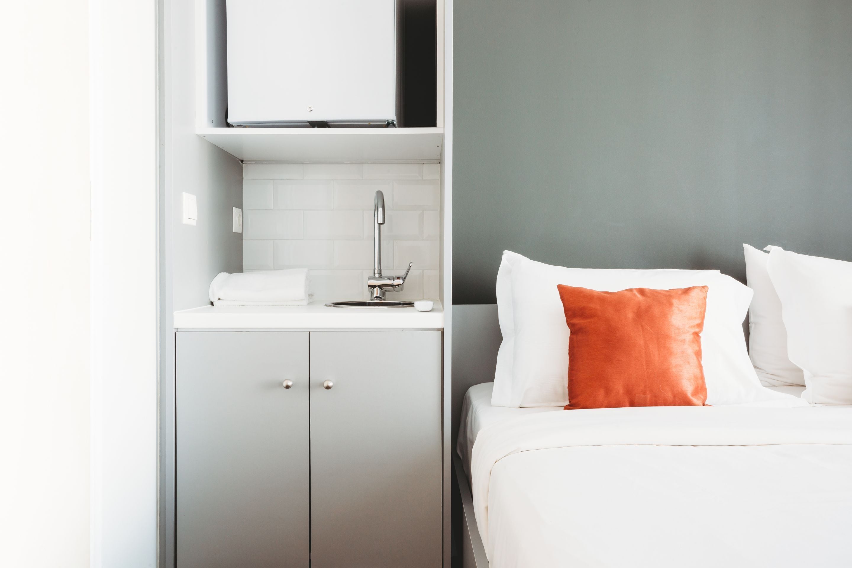 Una habitación de hotel con una cama, un armario gris, una pequeña cocina y una pared gris.