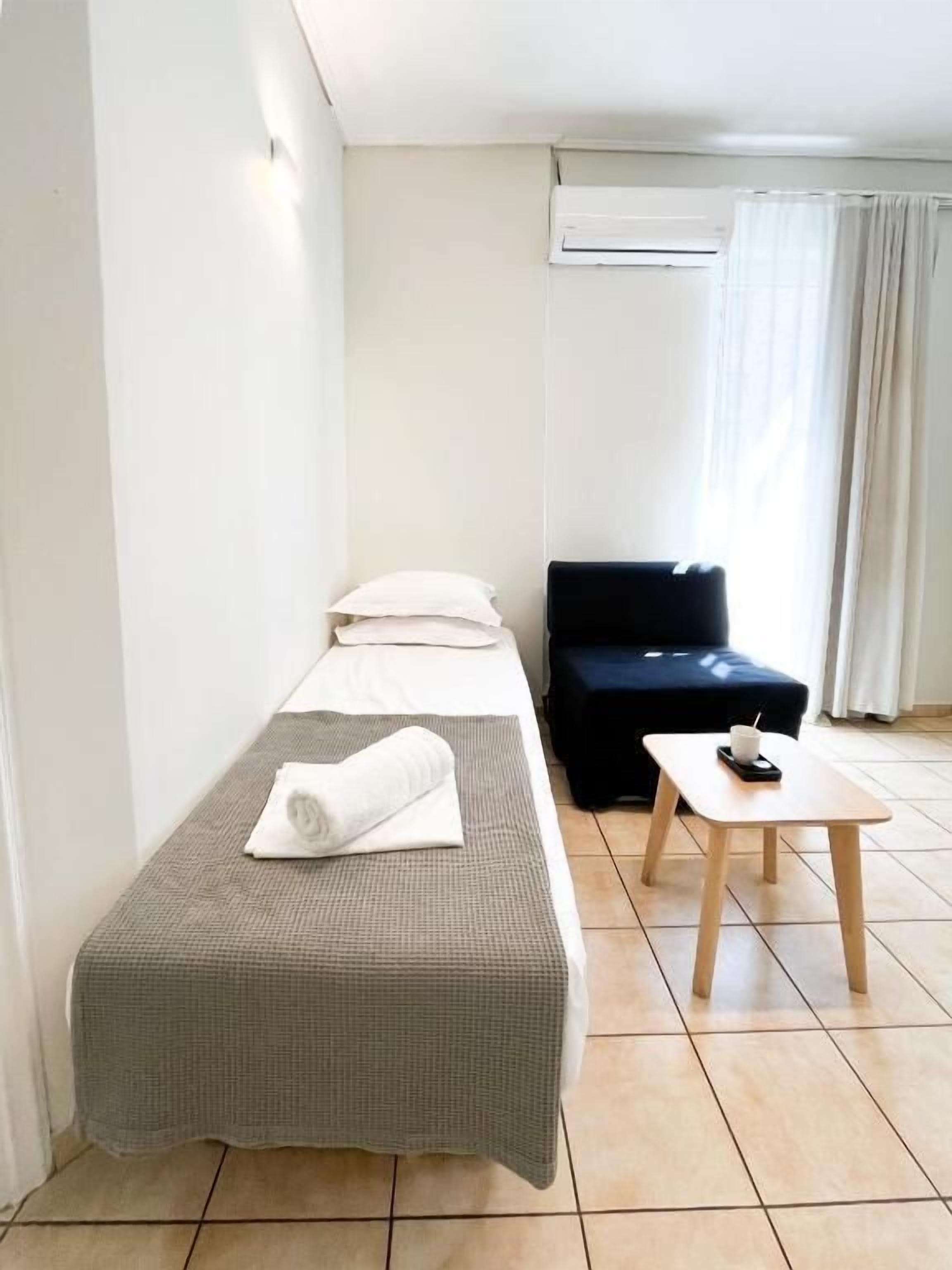 Una cama individual con una colcha gris de cuadros, una toalla blanca enrollada, un sofá negro, una mesita de madera con una taza y un platillo, y un equipo de aire acondicionado en la pared.