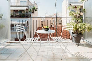 A balcony with a round table, two white chairs, a potted plant, and a bowl of fruit.