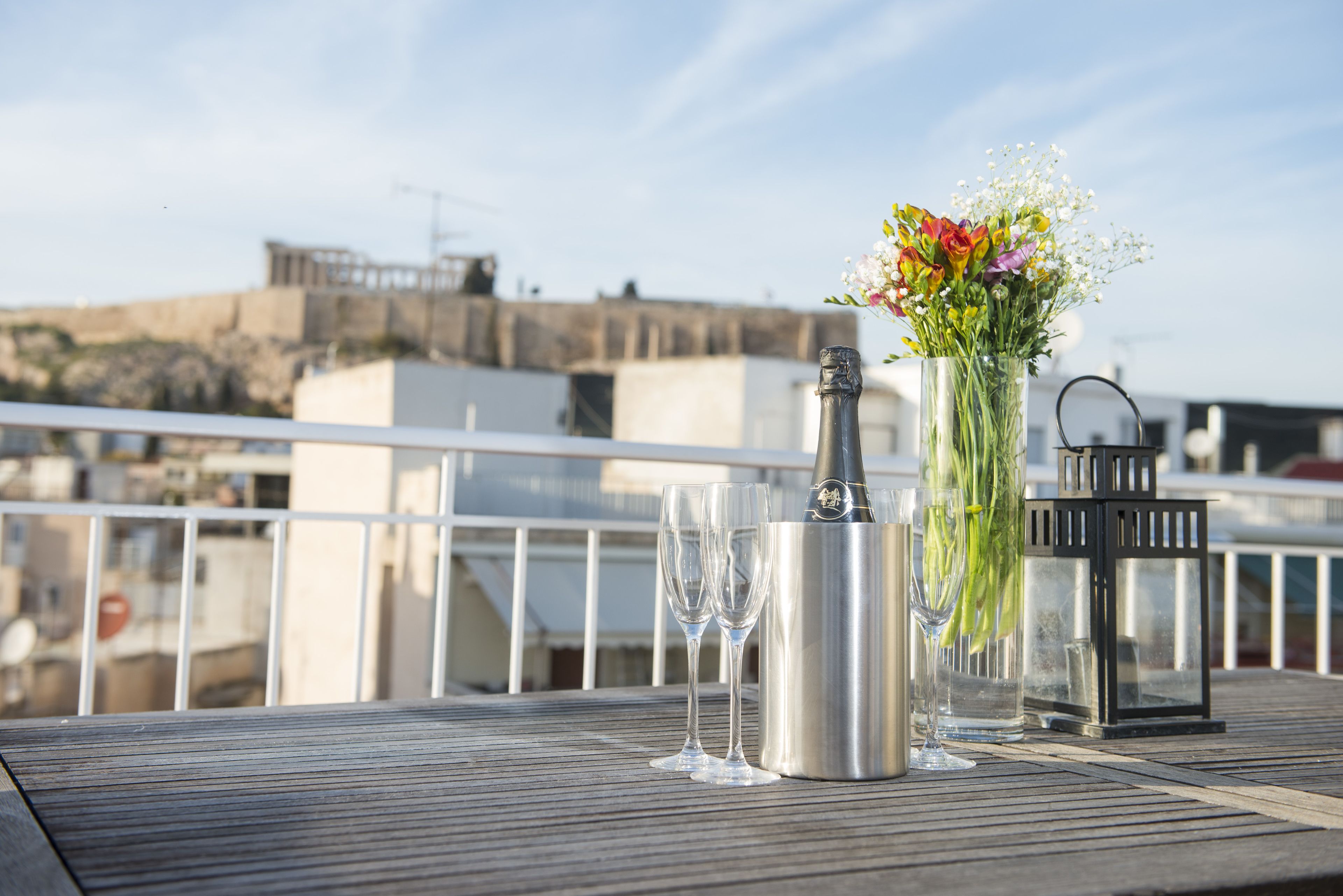 A table with a champagne bottle, two glasses, and a vase with flowers on a wooden deck with a railing.