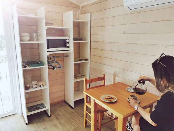 A compact kitchen with a microwave, shelves, and a dining table with a plate of cookies. - Camping Hameau Des Cannisses (Gruissan)