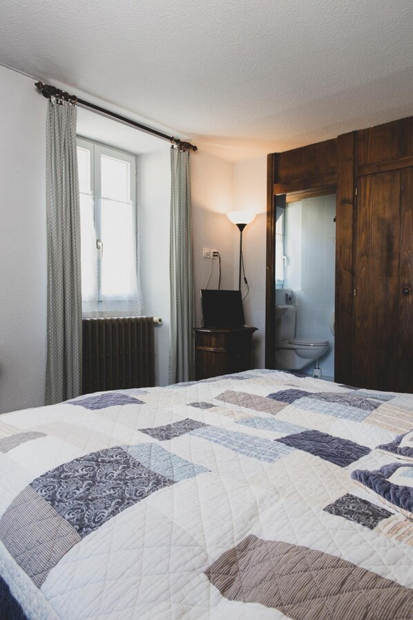 A bedroom with a large bed, a nightstand, a lamp, a wooden wardrobe, and a window with curtains. - Hotel Besso (Anniviers)