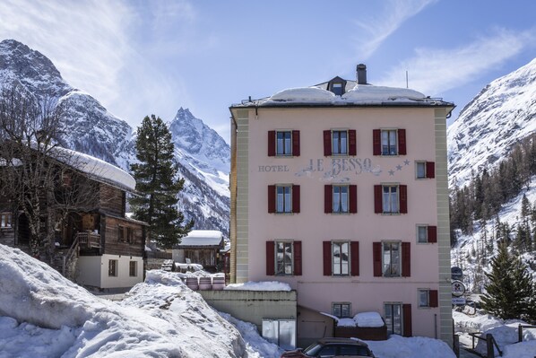 Un edificio rosa con diverse finestre e un tetto imbiancato dalla neve, con montagne sullo sfondo e circondato da neve.