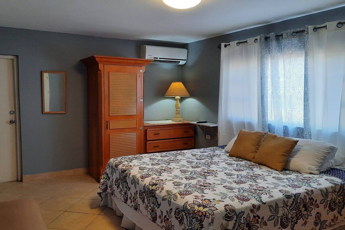 Un dormitorio con cama, un armario de madera, un escritorio con lámpara y una ventana con cortinas.