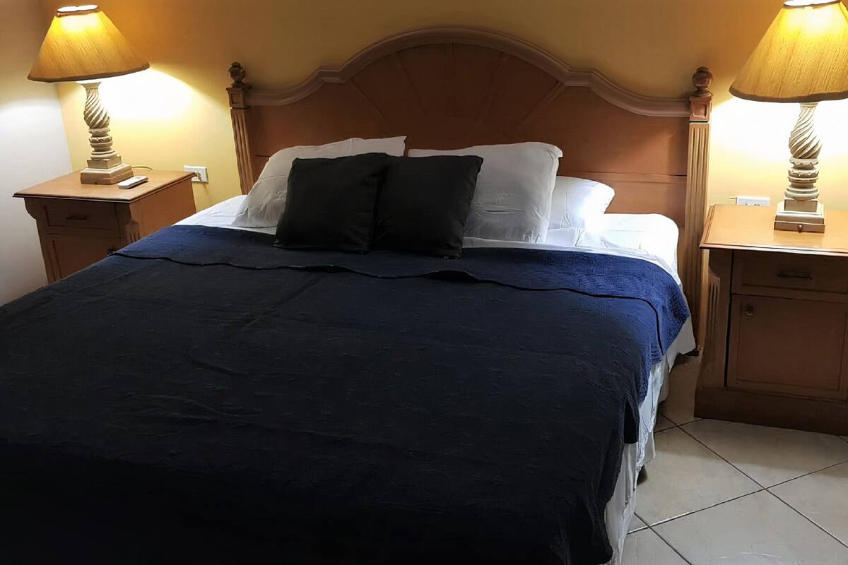 Una cama bien tendida con colcha azul y dos almohadas negras, flanqueada por dos mesitas de noche con lámparas, en una habitación con cabecera de madera y suelo de baldosas.