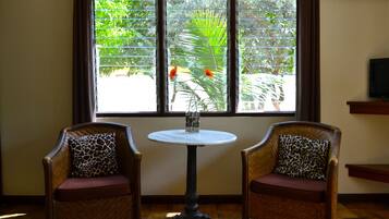 Quarto com duas cadeiras de vime com almofadas de estampa de leopardo, uma pequena mesa redonda e uma janela com vista para vegetação e uma palmeira.