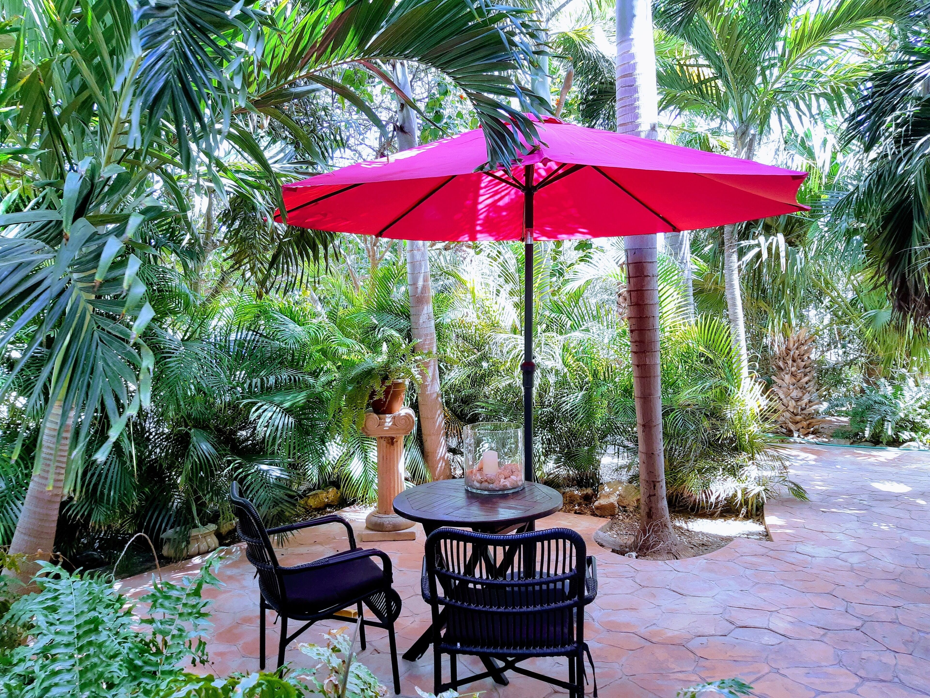 An outdoor patio with a round table and two chairs, a red umbrella, and lush tropical plants.