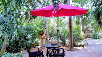 An outdoor patio with a round table and two chairs, a red umbrella, and lush tropical plants.