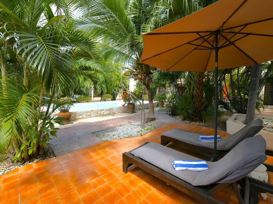 A poolside area with two lounge chairs under a large umbrella, surrounded by palm trees and a tiled floor.