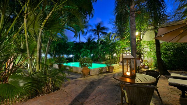 Un espacio al aire libre iluminado con luces de noche, que cuenta con una piscina, palmeras y un conjunto de mesa y sillas iluminadas por una linterna.