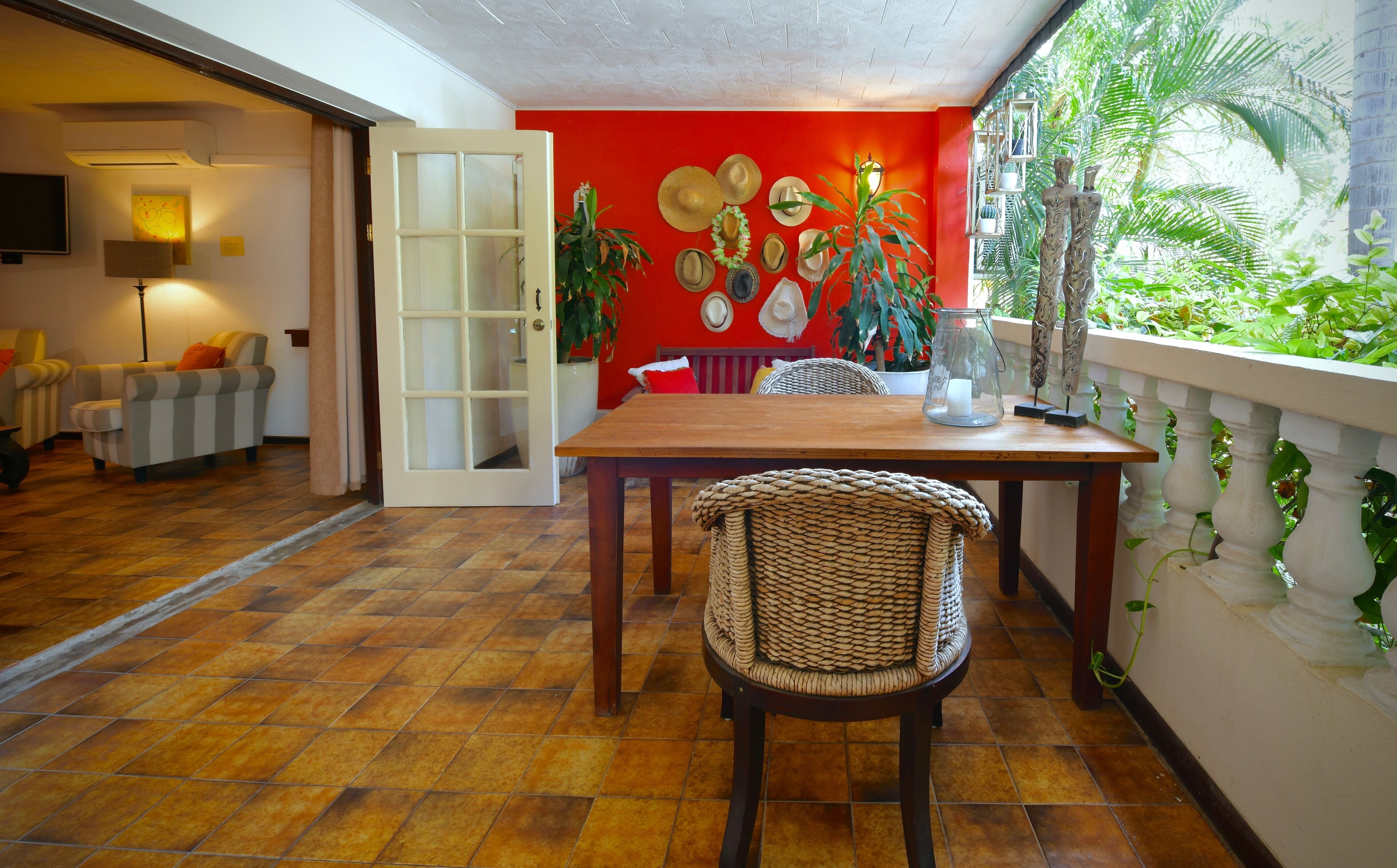 A dining area with a wooden table and wicker chair, a red accent wall, and a view of greenery through a balcony.